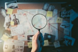 In the foreground is a hand holding a magnifying glass. In the blurry background a crimeboard full of picture, notes, and strings connect the dots and answering the question, What does a personal injury lawyer do in Hawaii?